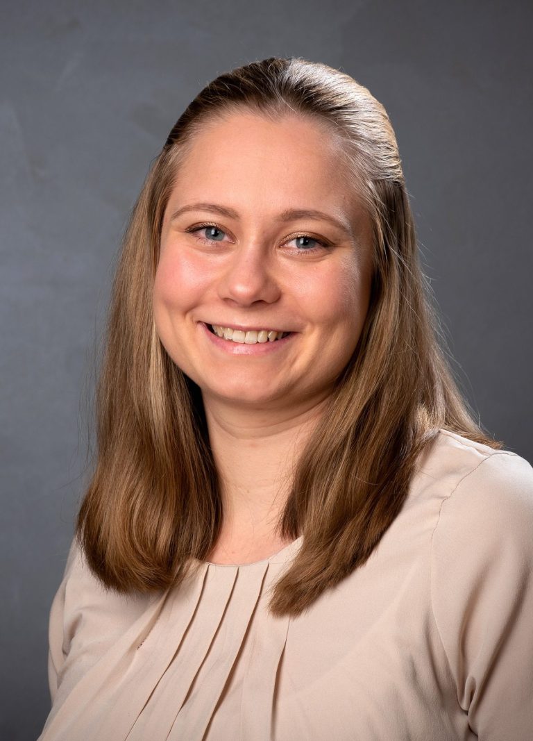 Lächelnde Person mit langen, blonden Haaren in hellem Oberteil vor grauem Hintergrund.